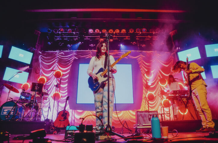 Lucy Dacus is pictured on stage, flanked by her band, at The Palace Theatre in Calgary Alberta. Behind her, screens displayed her "Home Video Tour" VHS logo can be seen. She is holding her guitar and singing.