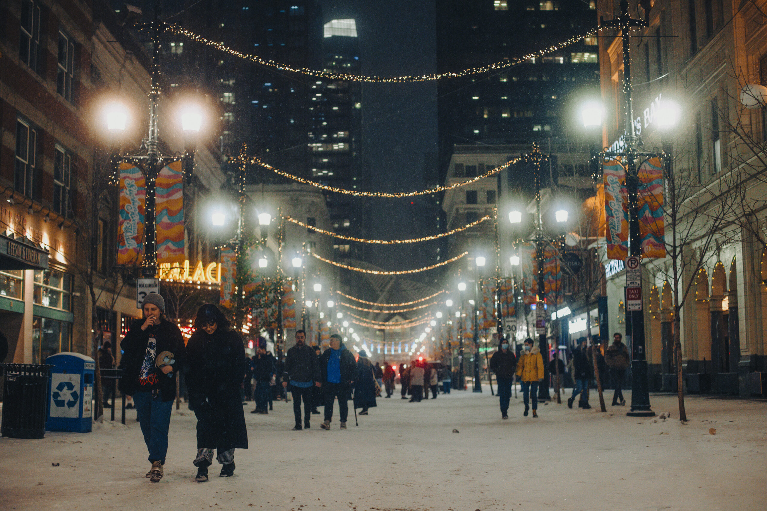 A snowy scene on Stephen Avenue in Calgary, Alberta as patrons walk to and from The Palace Theatre underneath hazy street lights.