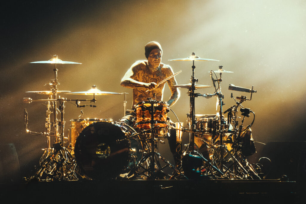 Travis Barker of Blink 182 is pictured onstage in golden light as he plays drums.
