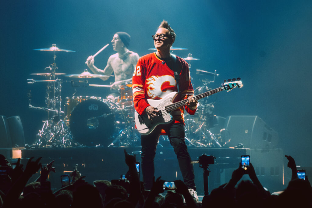 Mark Hoppus of Blink 182 is pictured on stage, standing and smiling while playing guitar.