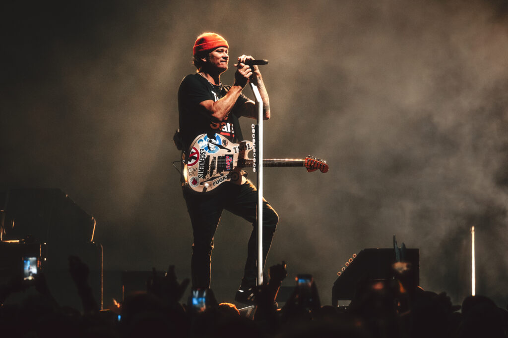 Blink 182's Tom DeLonge is pictured standing and smiling on stage as he grips the microphone.